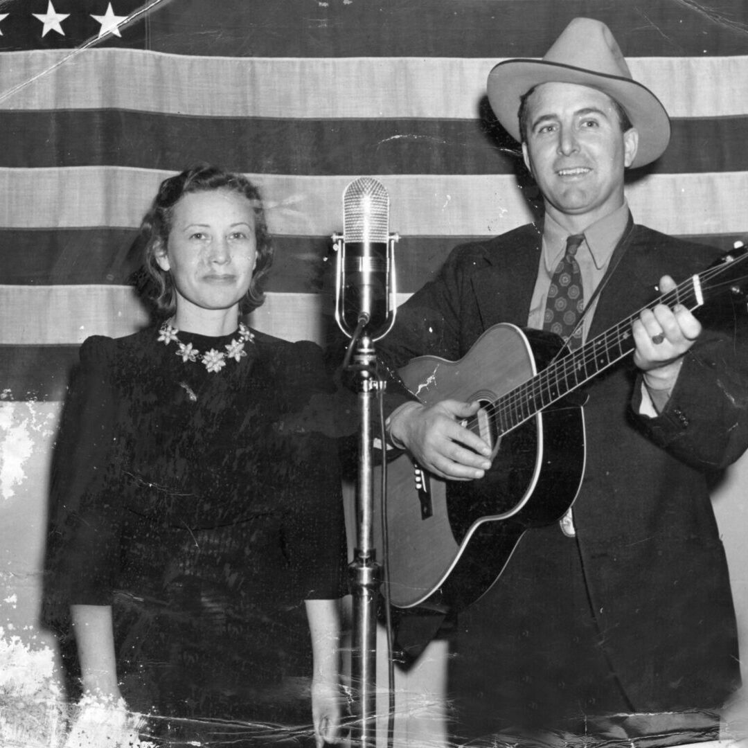 A man and woman sing into microphones in front of an american flag.